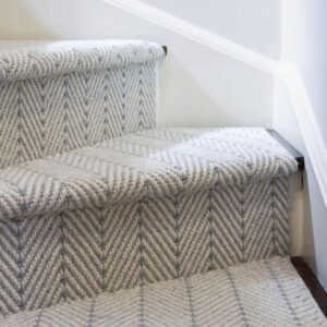 Light gray herringbone-patterned carpet on wooden stairs with white walls, showing modern home interior flooring design