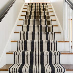 A staircase with white risers, wooden treads, and a black and white striped stair runner installed down the center.