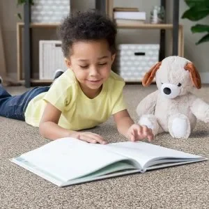 A young child lies on their stomach on a carpeted floor, engrossed in reading an open book, with a plush dog toy sitting beside them