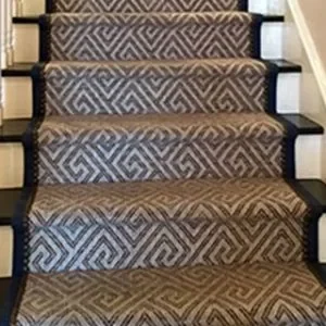 A close-up view of a staircase featuring a patterned stair runner with a repeating geometric design, possibly in shades of brown and beige, contrasting with dark-colored stair treads and risers.
